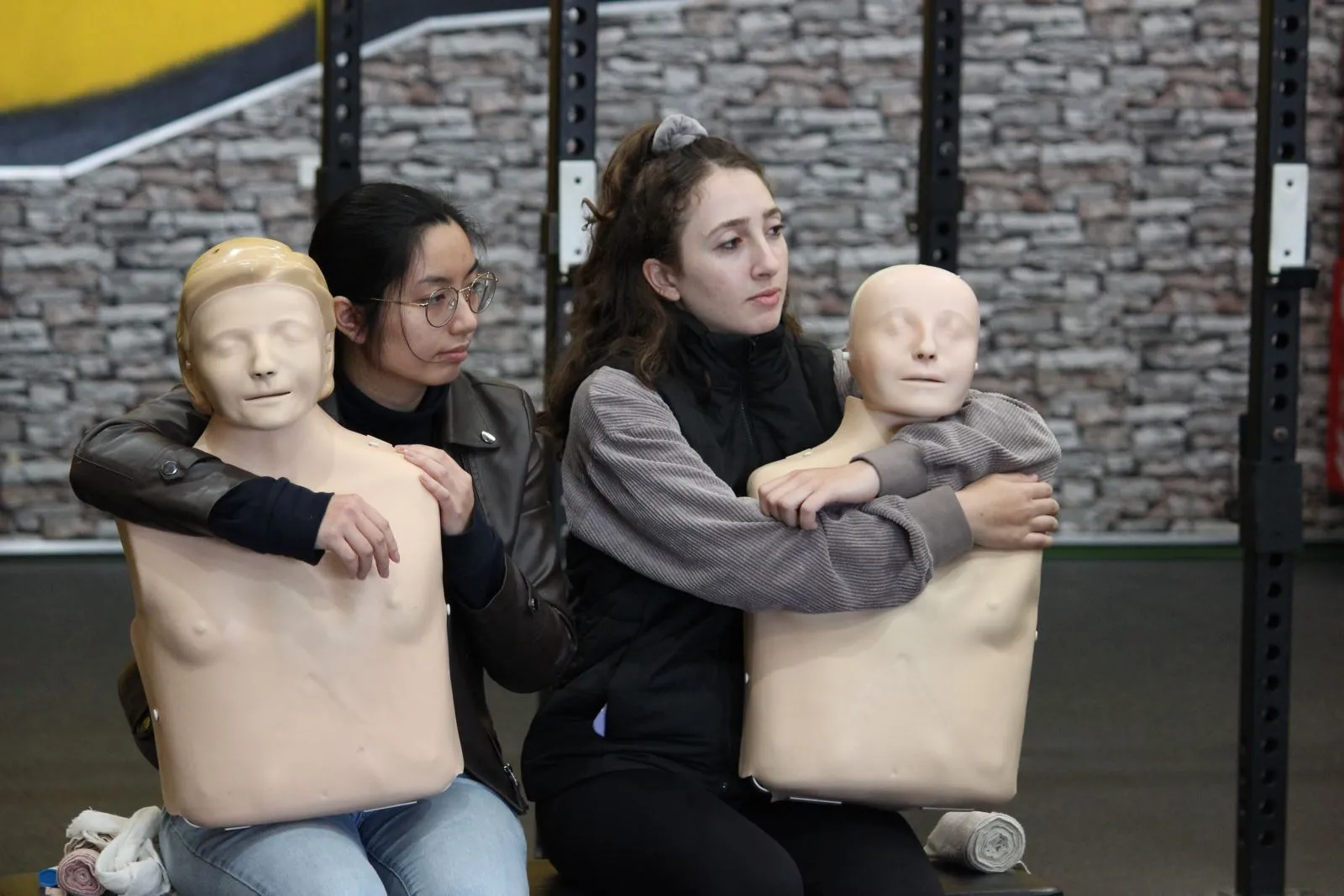 Two young women sit with CPR mannequins in a training setting.
