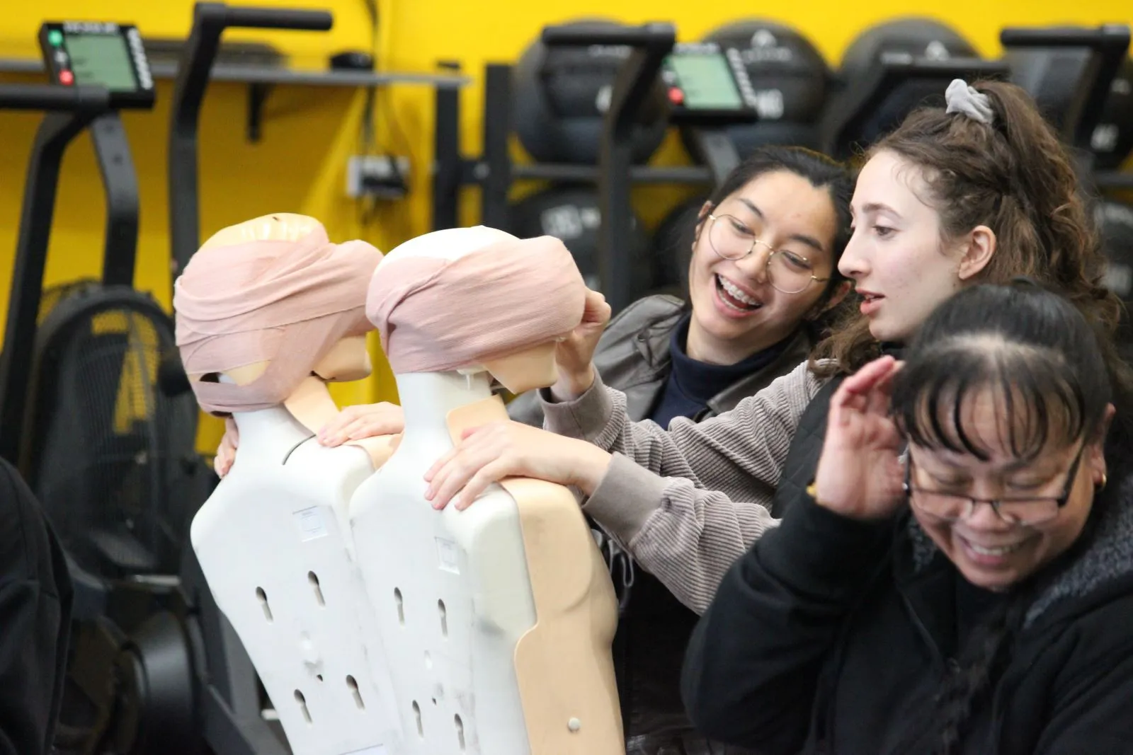 Two people adjust bandages on mannequins in a gym setting.