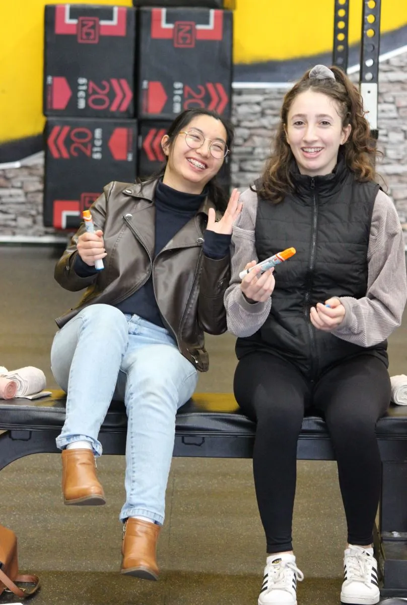 Two smiling friends sitting together with markers in a gym.