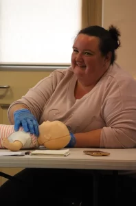A woman practices CPR on a baby doll while smiling in a classroom.