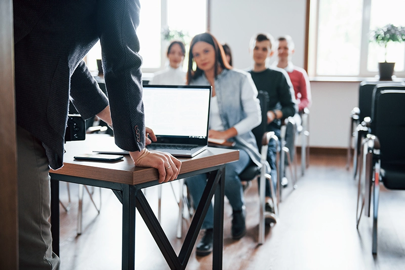 A speaker stands at a table in front of an audience during a presentation.